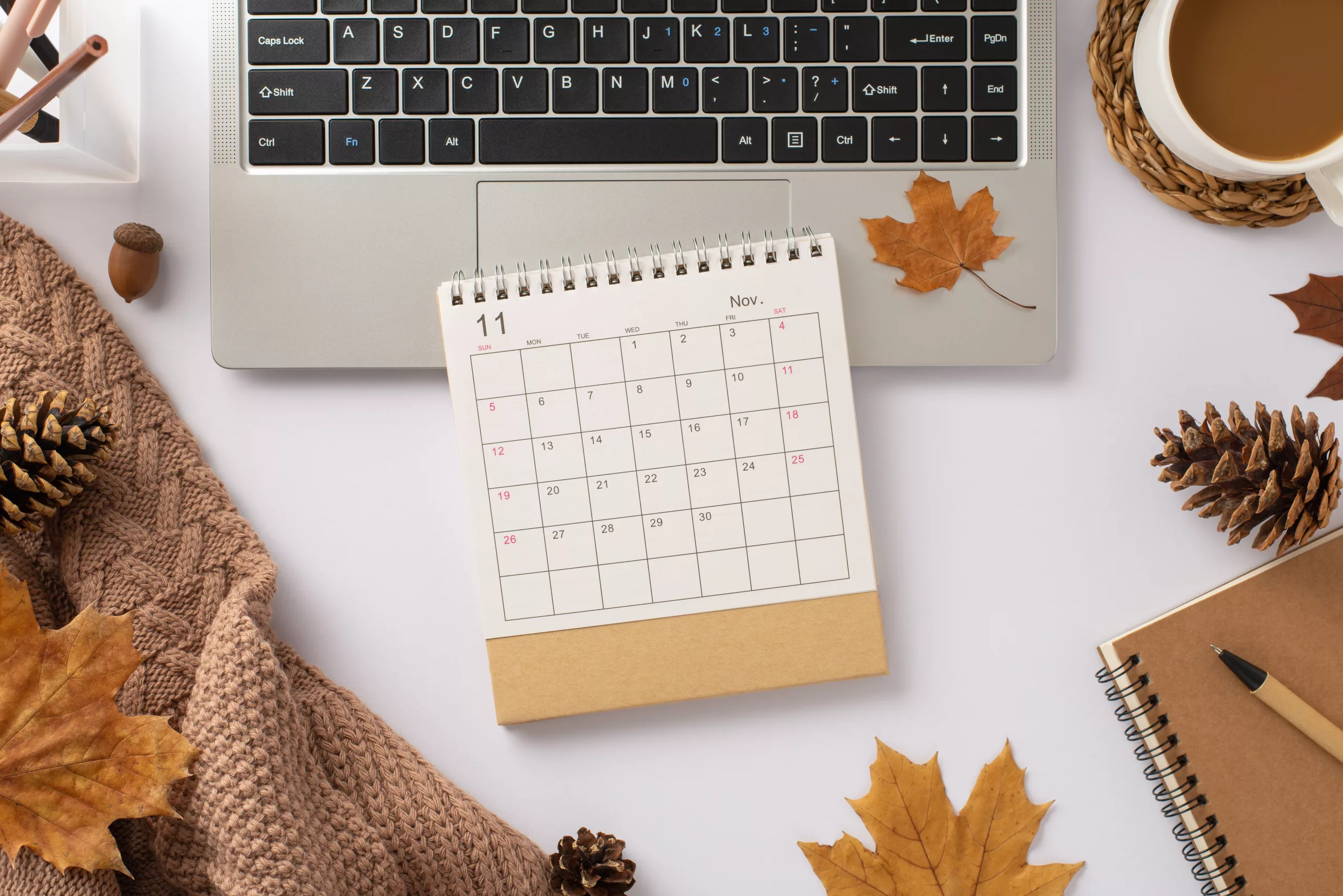 Productivity meets autumn comfort. Top view: laptop, cozy plaid, hot cocoa, spiral notepad, pen, calendar, pine cones, maple leaves. Curate your fall-inspired workspace